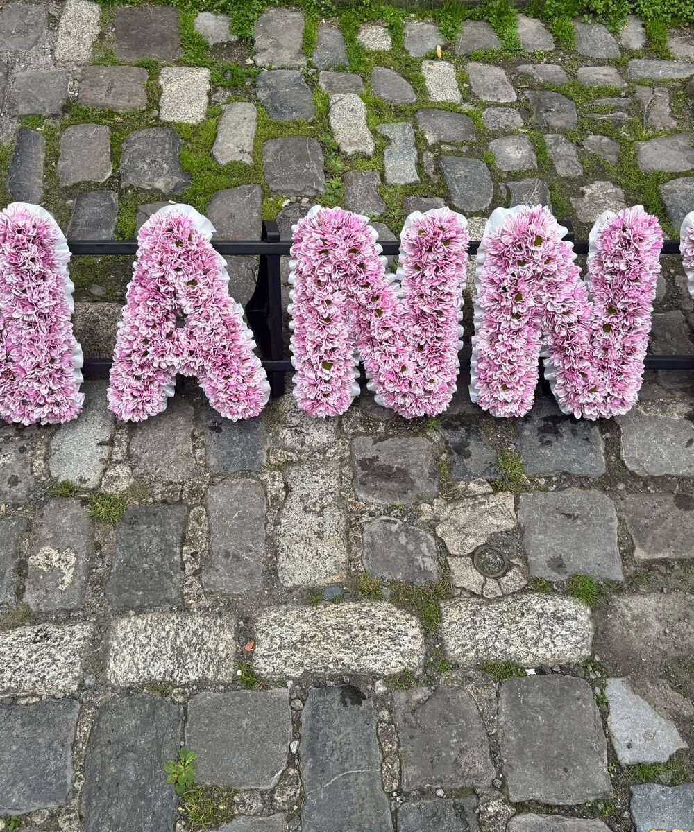 NANNY lettered funeral tribute in pink chrysanthemum with pastel floral corner sprays, by Bristol Floral Design