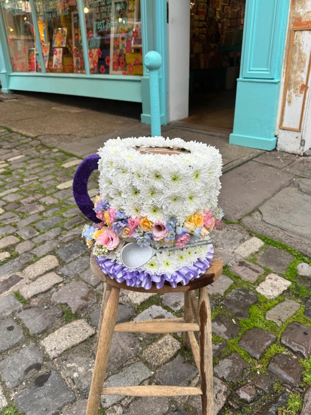 Teacup and saucer funeral tribute in white chrysanthemum with pastel garden flowers and a silver spoon, by Bristol Floral Design